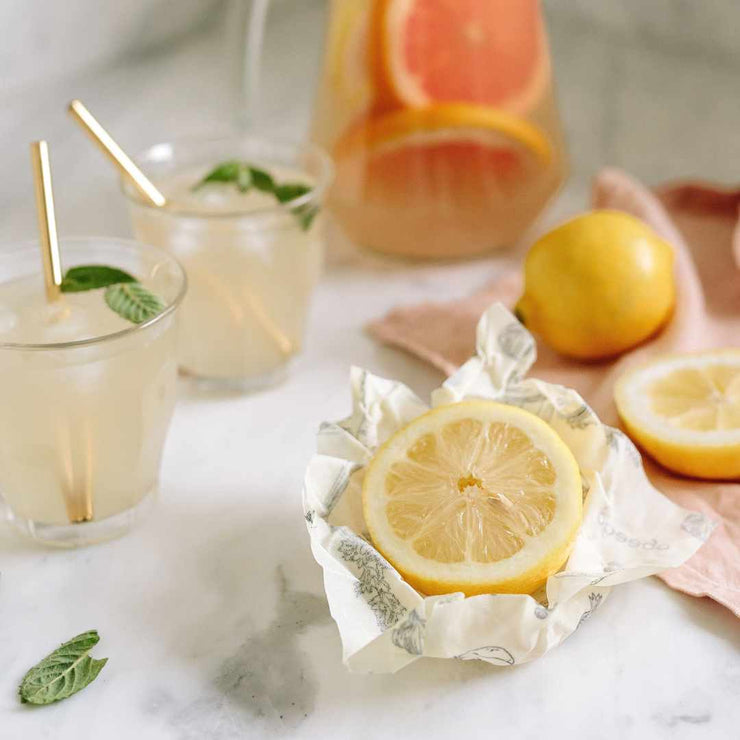 Half a lemon partially wrapped in a small Abeego beeswax food wrap surrounded by freshly made cocktails.